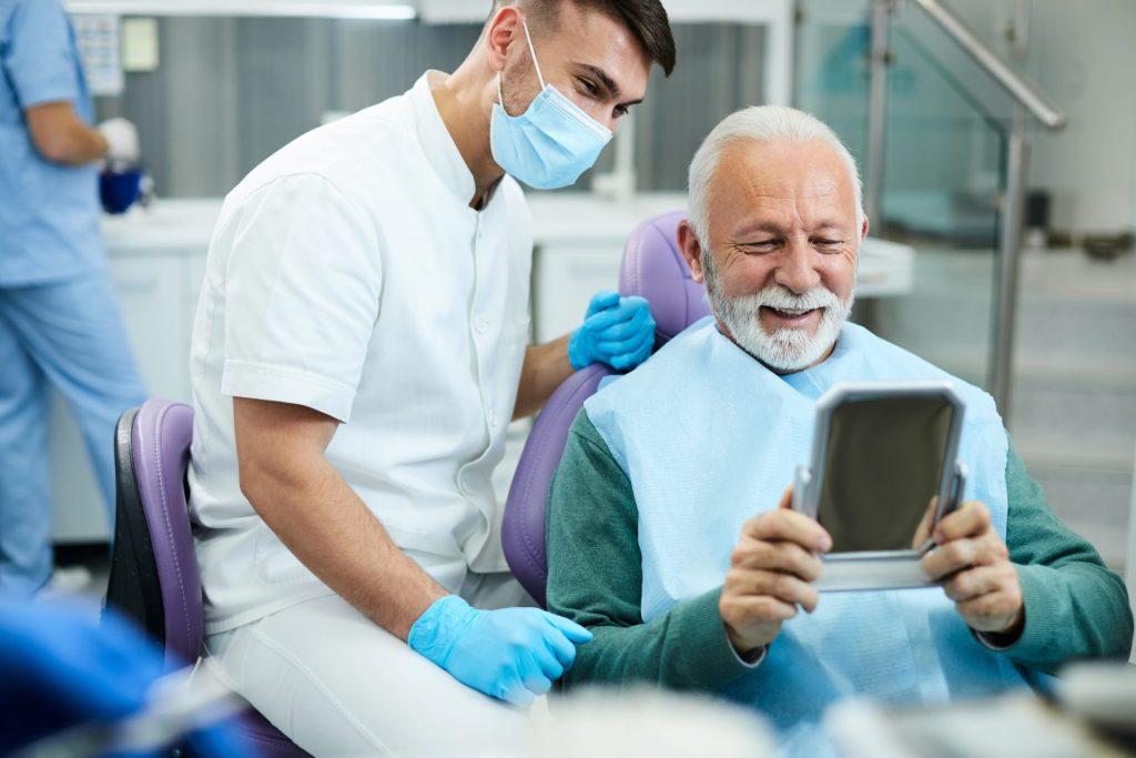 Smiling senior man checking his new teeth in a mirror with his dentist