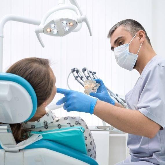 Dentist in mask and gloves discusses dental model with patient in dental chair under overhead light